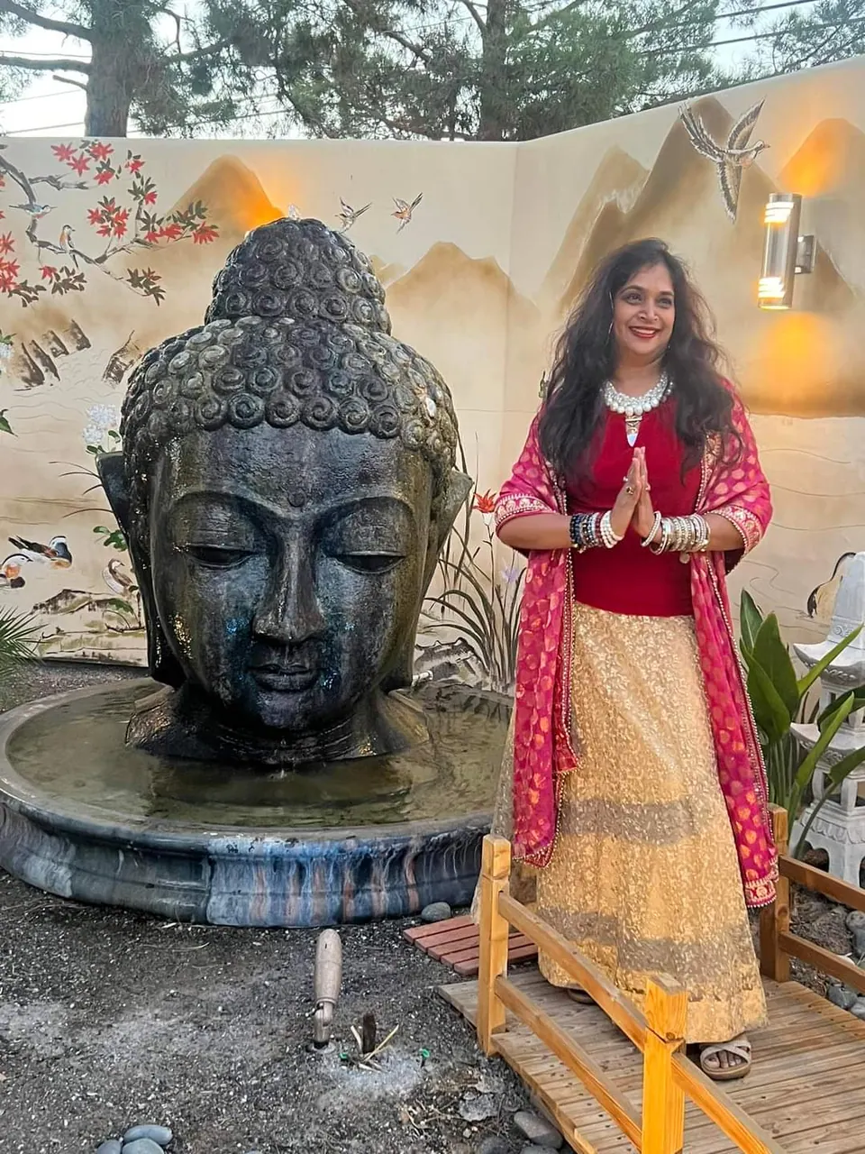 Bhavna with her hands together in greeting, beside a large stone Buddha head