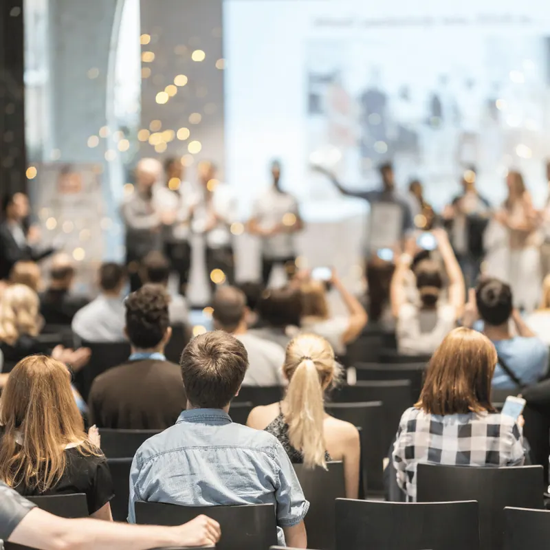 An audience applauding with hands raised in front of a lit stage
