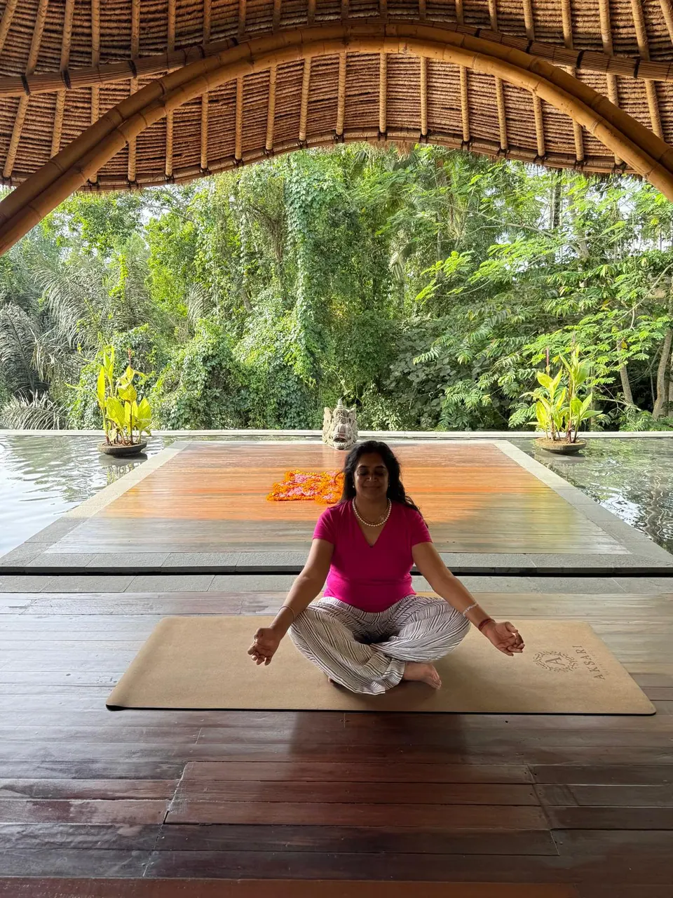 A woman meditating cross-legged on a wooden deck in an open-air pavilion surrounded by tropical trees