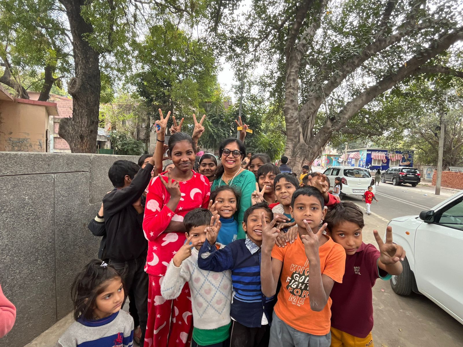 A group of smiling children gathered around a woman on a tree-lined street, waving at the camera