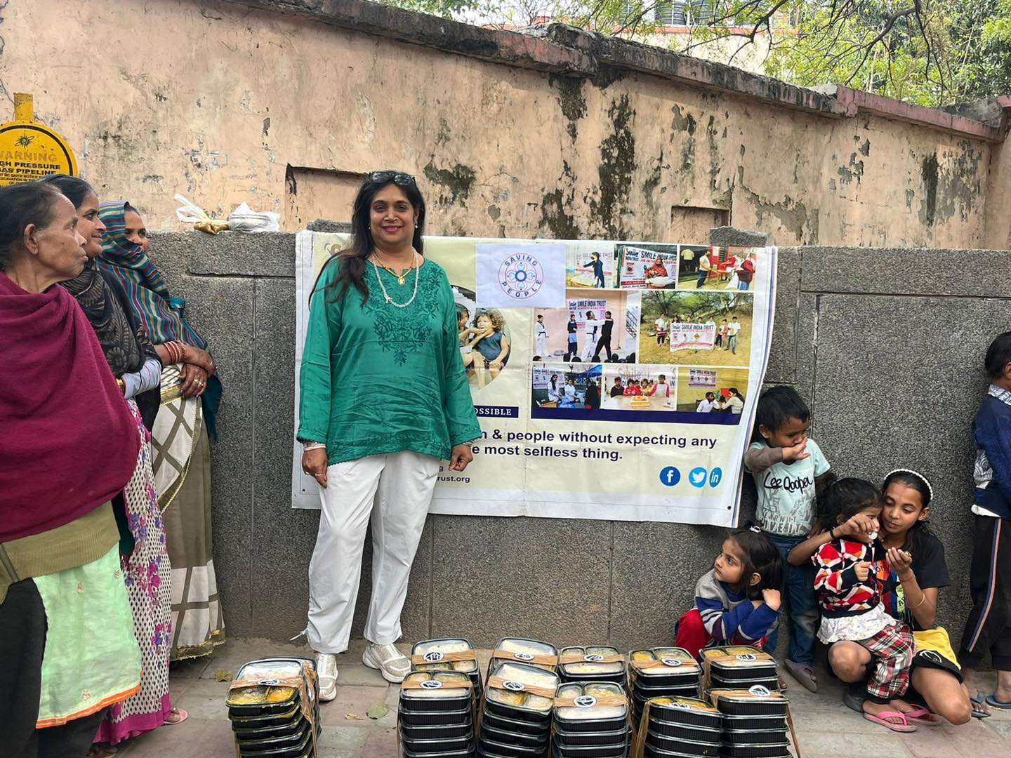Bhavna standing beside a Saving People banner, with rows of packed meals on the ground and local families gathered around her