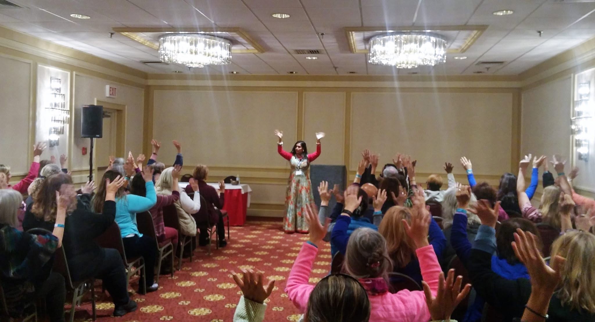 A room of women with hands raised, following a speaker in white at a hotel ballroom event