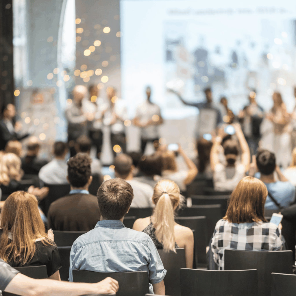 An audience applauding with hands raised in front of a lit stage