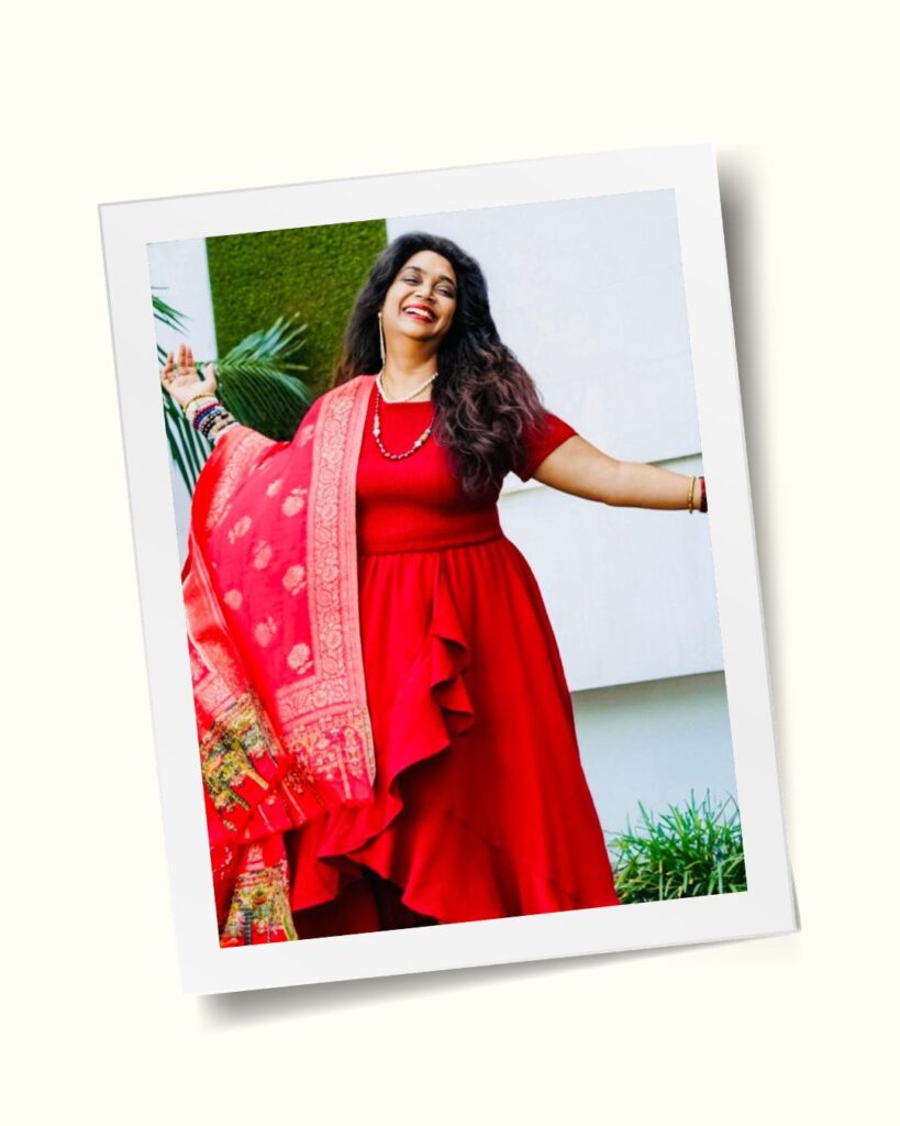 Bhavna smiling with her arms open, in a red dress and pink embroidered scarf, in front of a wall of green foliage