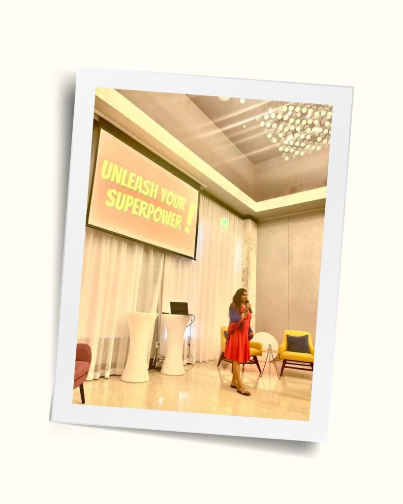 Bhavna speaking on stage in a red dress, beside a screen reading Unleash Your Superpower
